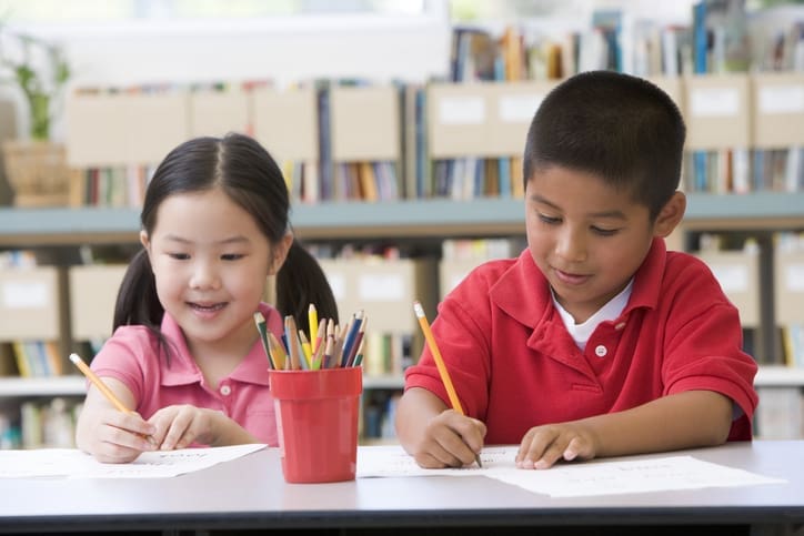 Kindergarten children sitting at desk and writing in classroom holding pencils